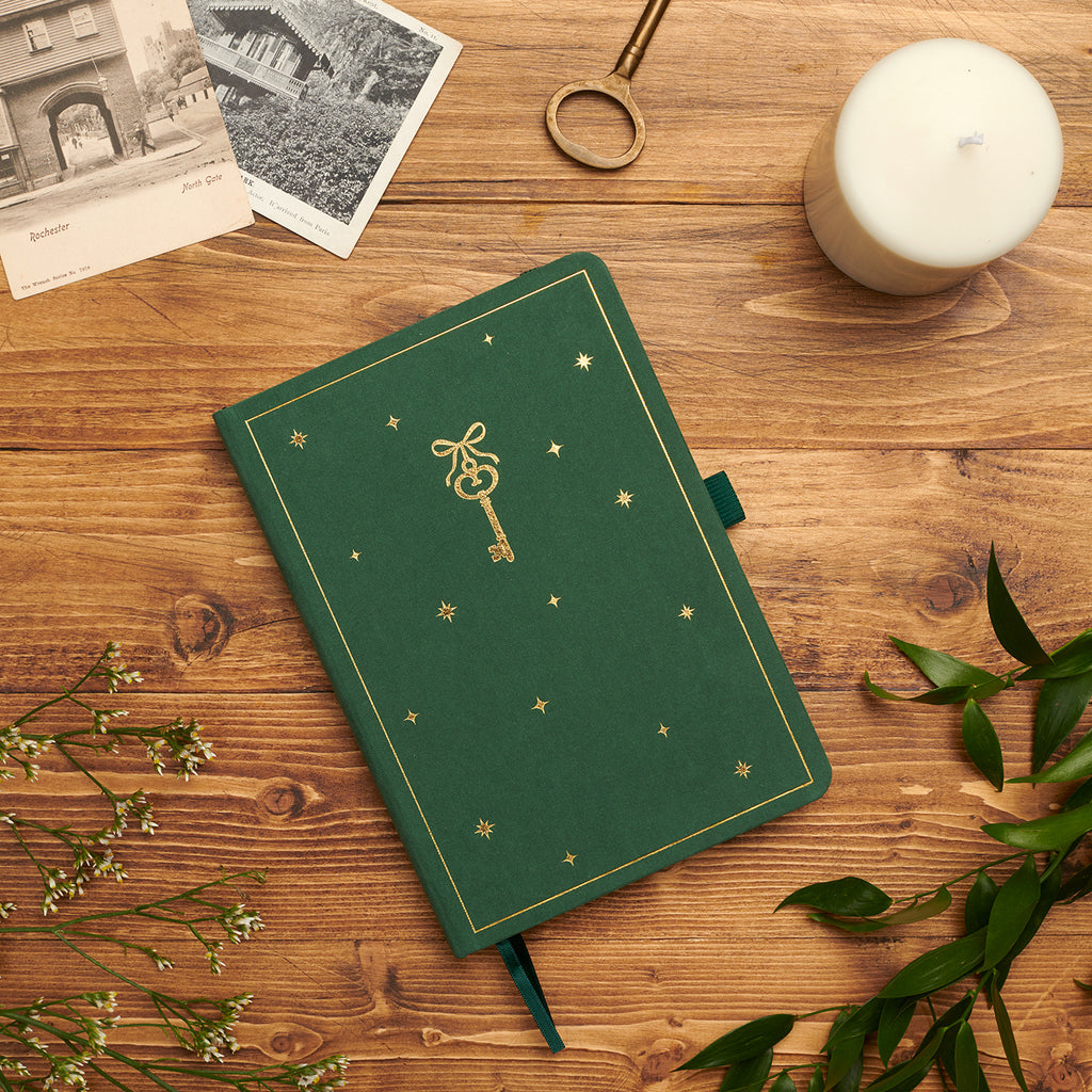 Enchanted Key dotted notebook — green velvet cover with gold-foil key motif on wooden desk beside candle and ribbon.
