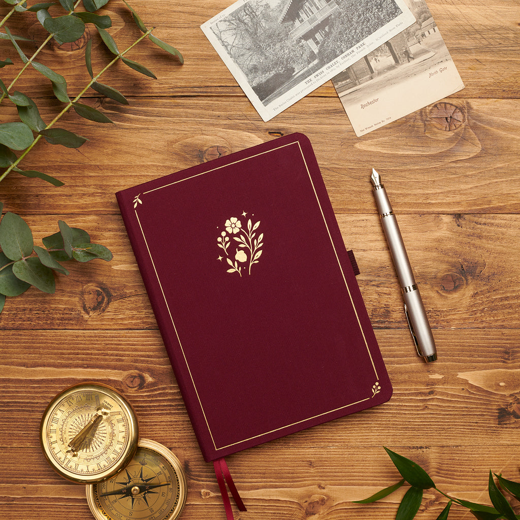 Plum linen journal with gold floral motif styled with a vintage compass, postcards, and pen.