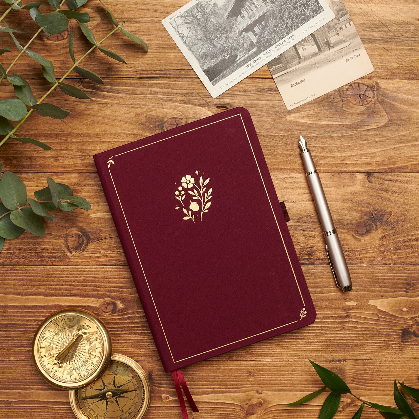 Plum linen journal with gold floral motif styled with a vintage compass, postcards, and pen.
